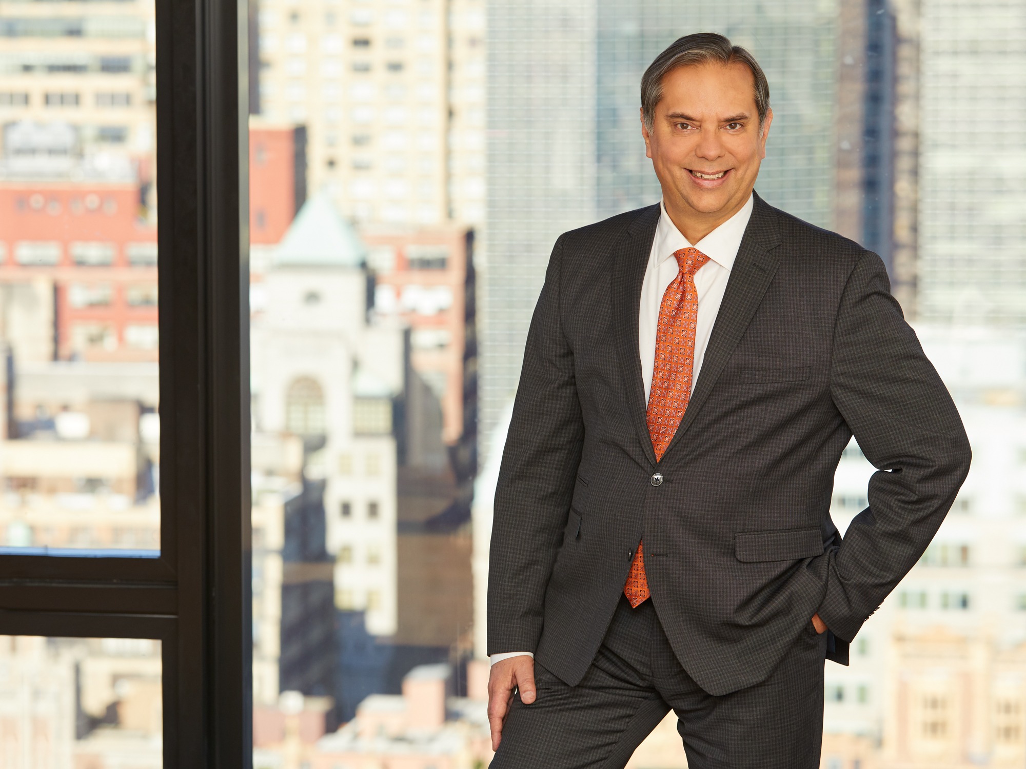 Professional man in a suit smiles confidently with a city skyline in the background, illustrating leadership and success in a corporate environment.